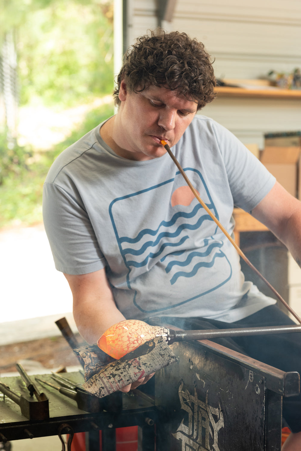 Brad smith seated at his glass blowing bench with a wet newsprint shaping a globe of hot glass.