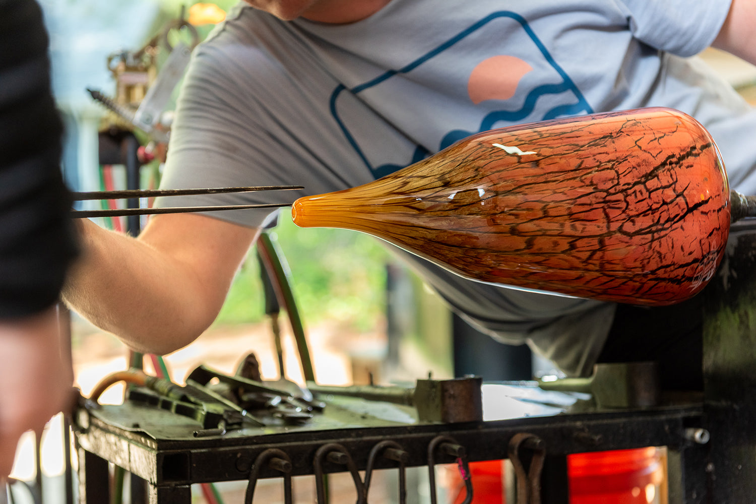 An in-progress hot glass vase on it's side against the background of glass artist Brad Smith, who's tools are poised to work in it